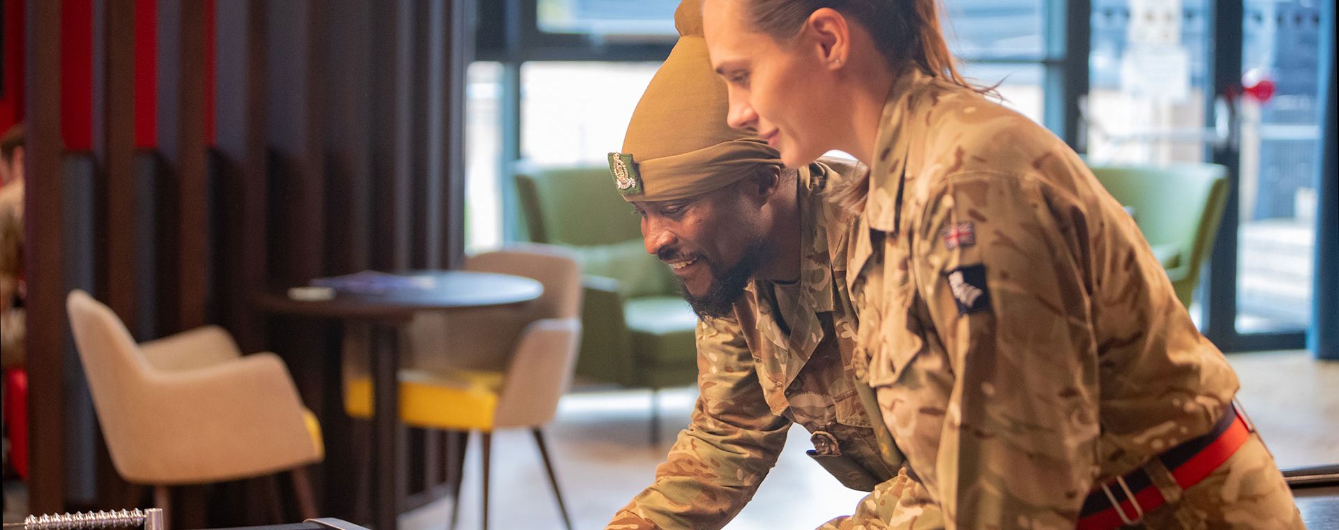 Two soldiers in uniform play foosball together, smiling and focused in a recreational room with chairs and a table in the background.