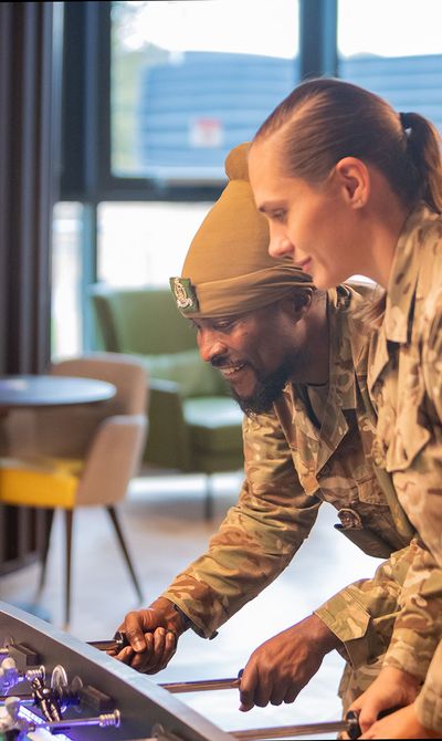 Two soldiers in uniform play foosball together, smiling and focused in a recreational room with chairs and a table in the background.