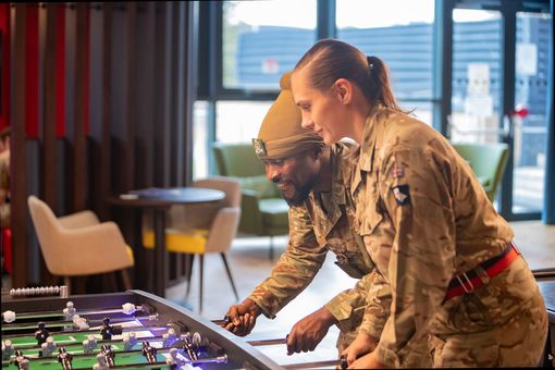 Two soldiers in uniform play foosball together, smiling and focused in a recreational room with chairs and a table in the background.
