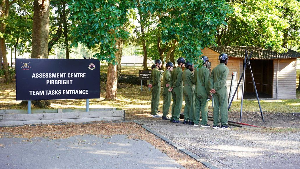 A group of people in green uniforms stand in line near a sign for the "Assessment Centre Pirbright" in a wooded area.