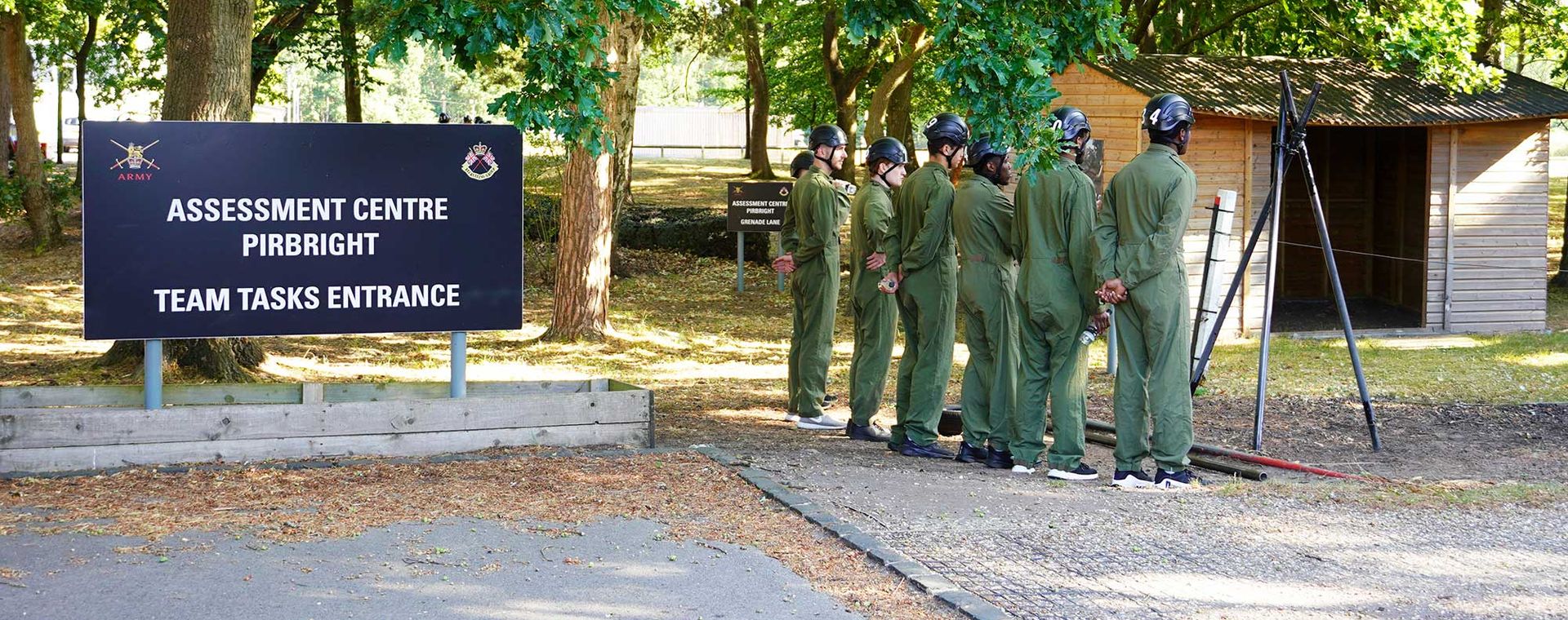 A group of people in green uniforms stand in line near a sign for the "Assessment Centre Pirbright" in a wooded area.