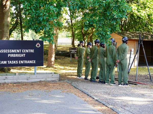 A group of people in green uniforms stand in line near a sign for the "Assessment Centre Pirbright" in a wooded area.