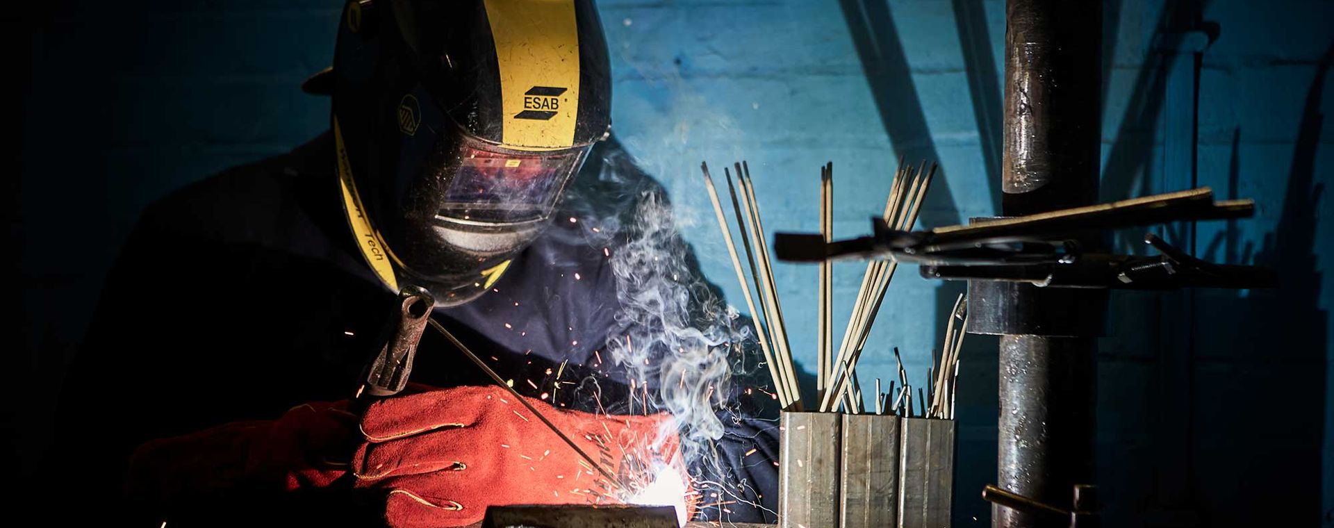 Welder in protective gear works with a torch, surrounded by metal rods and smoke, in a dimly lit workshop.