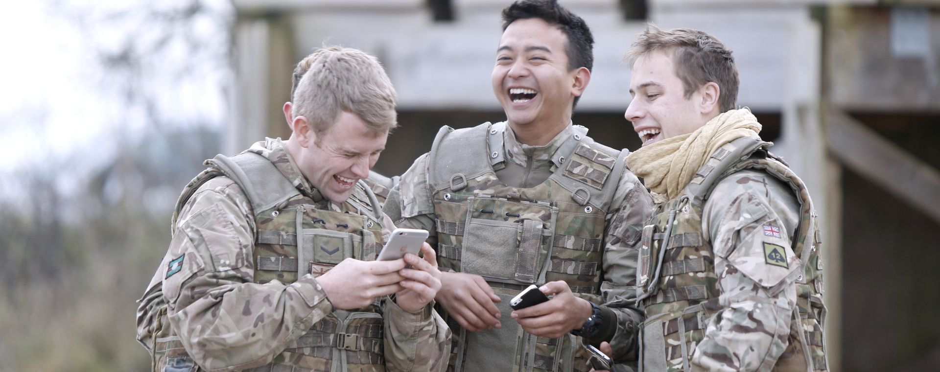 Three soldiers in camouflage uniforms laughing and holding phones outdoors