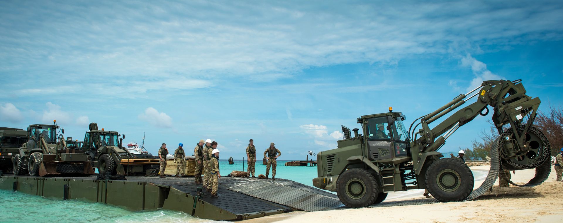 Military personnel and vehicles on a beach, using a temporary bridge to unload equipment from the sea under a clear blue sky.
