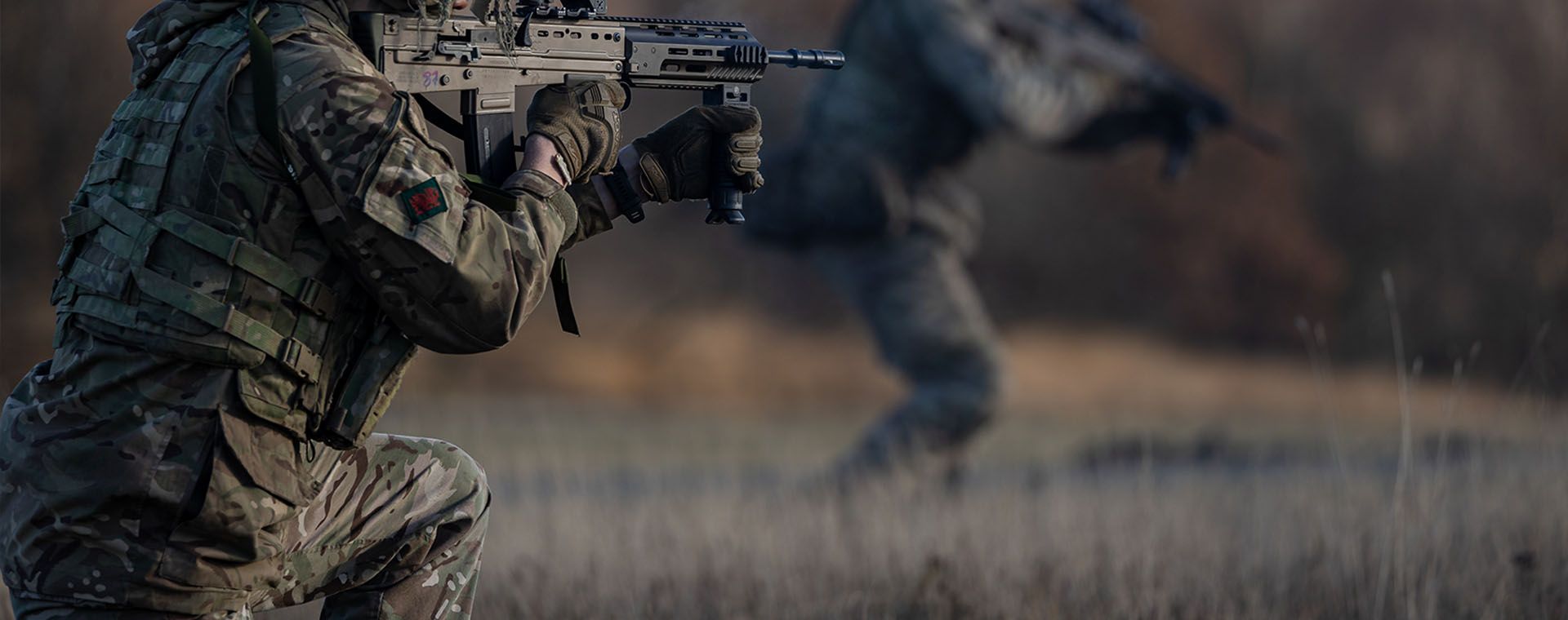 Two soldiers in camouflage gear, one kneeling with a rifle, the other moving forward in a blurred background, with autumn trees behind.