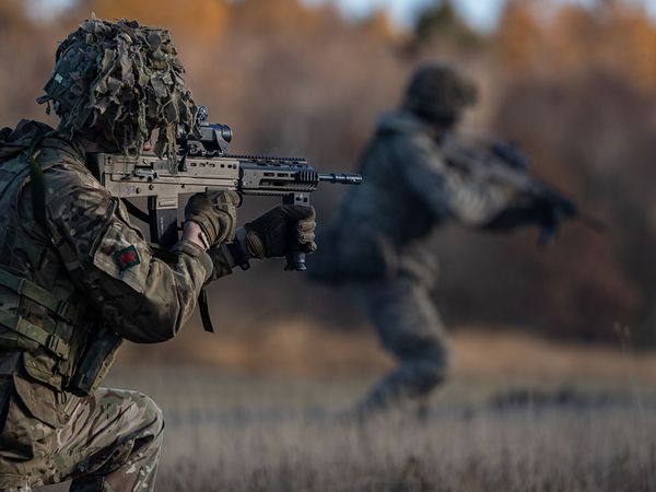 Two soldiers in camouflage gear, one kneeling with a rifle, the other moving forward in a blurred background, with autumn trees behind.