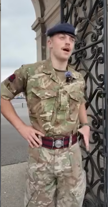 A soldier in camouflage uniform and beret stands at ease near an ornate wrought iron gate, with a building and cloudy sky in the background.