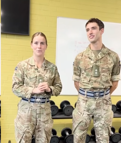 Two military personnel in uniform stand in a room with a yellow brick wall and dumbbells in the background.