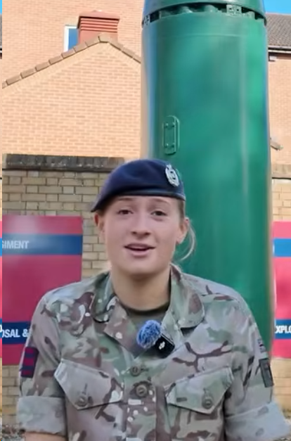 A person in a military uniform stands in front of a green object, outdoors, with a brick building in the background.