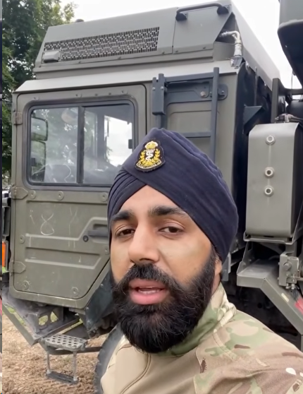 A man in a military uniform and turban stands in front of a green military vehicle outdoors.