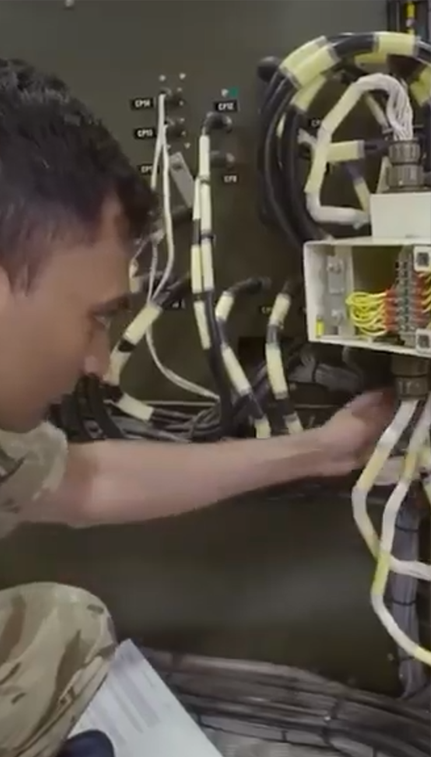 Man in uniform working on electrical wiring, connecting cables in a control panel, focused on task with various colored wires visible.