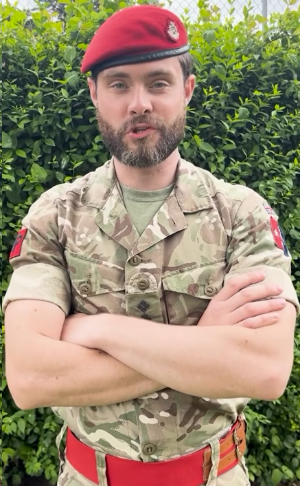 A man in a camouflage military uniform and red beret stands with arms crossed in front of a leafy background.