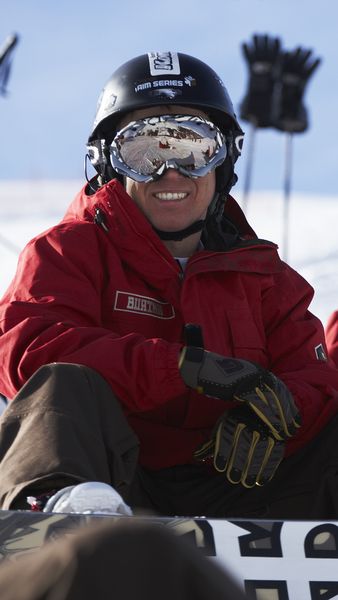 Three snowboarders in red jackets and helmets sit on the snow, smiling, with a snowy landscape in the background.