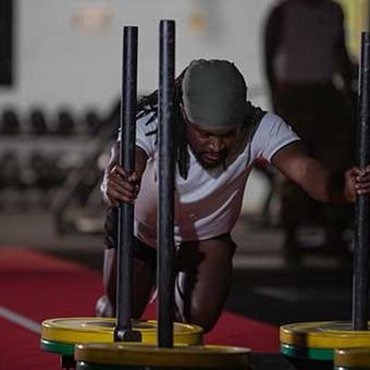 Man in a gym pushing a weighted sled on a red track, wearing a white shirt and cap, with gym equipment in the background.
