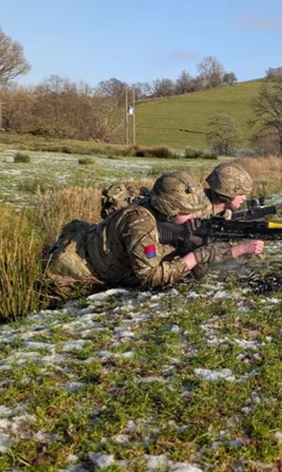 Soldiers from the Royal Artillery in camouflage gear are lying prone on snowy grass, aiming rifles, while an instructor stands behind them in a field.