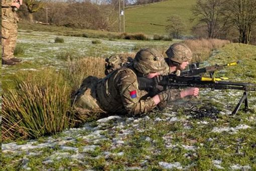 Soldiers from the Royal Artillery in camouflage gear are lying prone on snowy grass, aiming rifles, while an instructor stands behind them in a field.