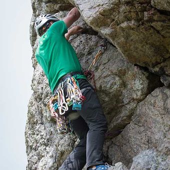 A climber in a green shirt and helmet ascends a rocky cliff, equipped with climbing gear and ropes.