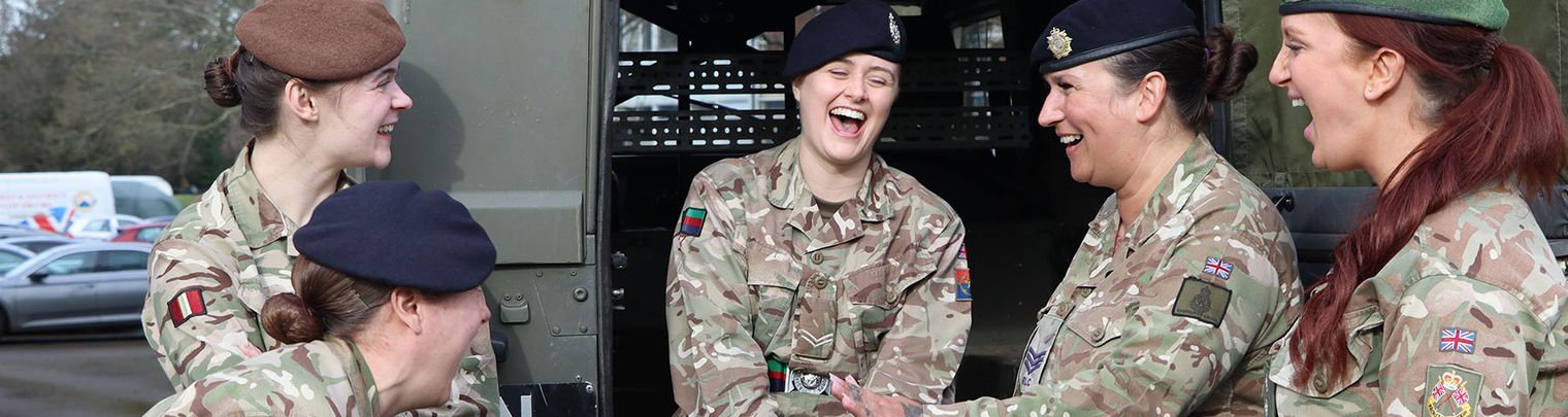 Five soldiers in camouflage uniforms sit and stand by a military vehicle, laughing and enjoying each other's company.
