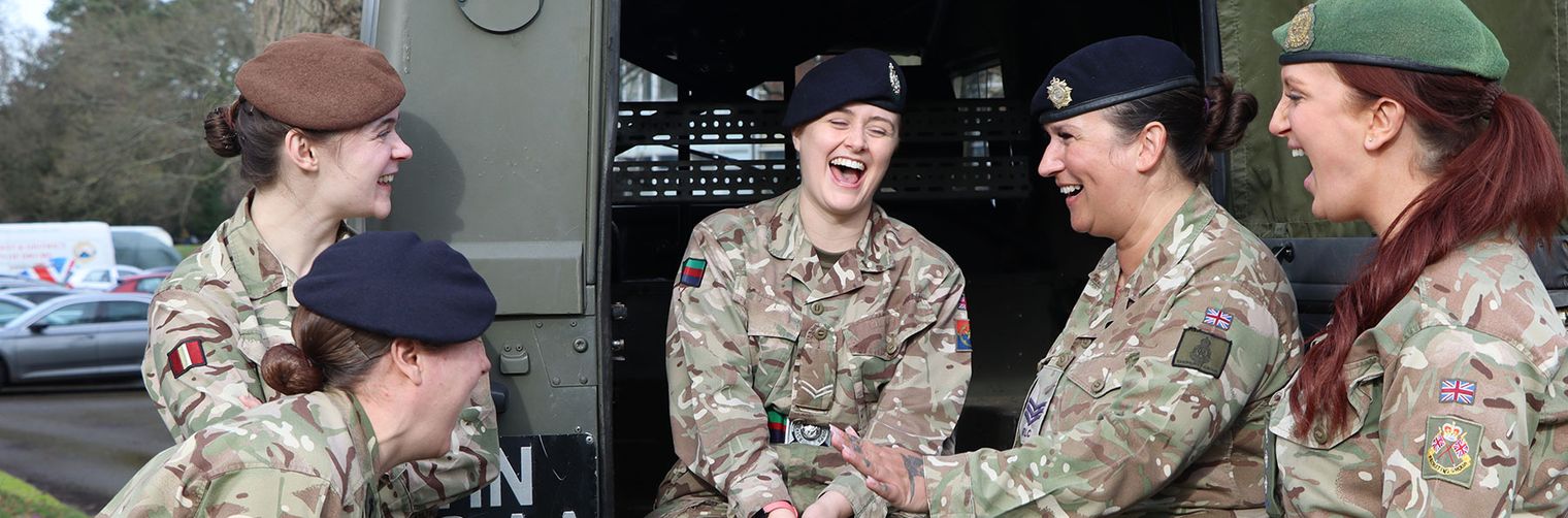 Five soldiers in camouflage uniforms sit and stand by a military vehicle, laughing and enjoying each other's company.