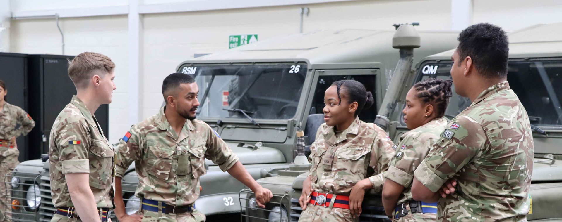 Five soldiers in camouflage uniforms stand and converse by military vehicles in a garage setting.