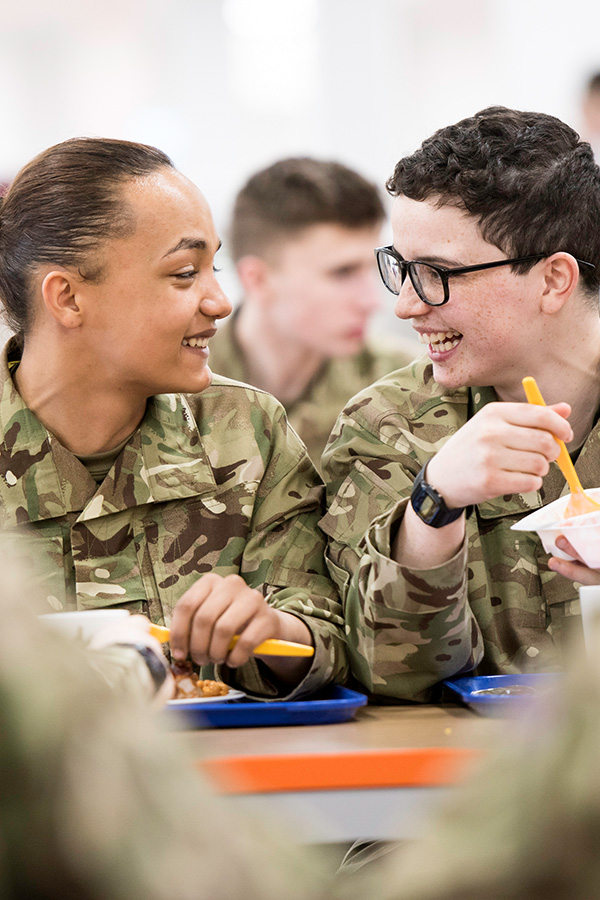 Two individuals in camouflage uniforms smile and converse while eating at a cafeteria table.