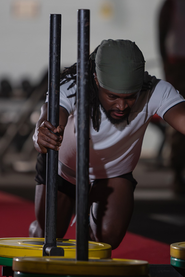 Man in a gym pushing a weighted sled, wearing a green headband and white shirt, focused on his workout.