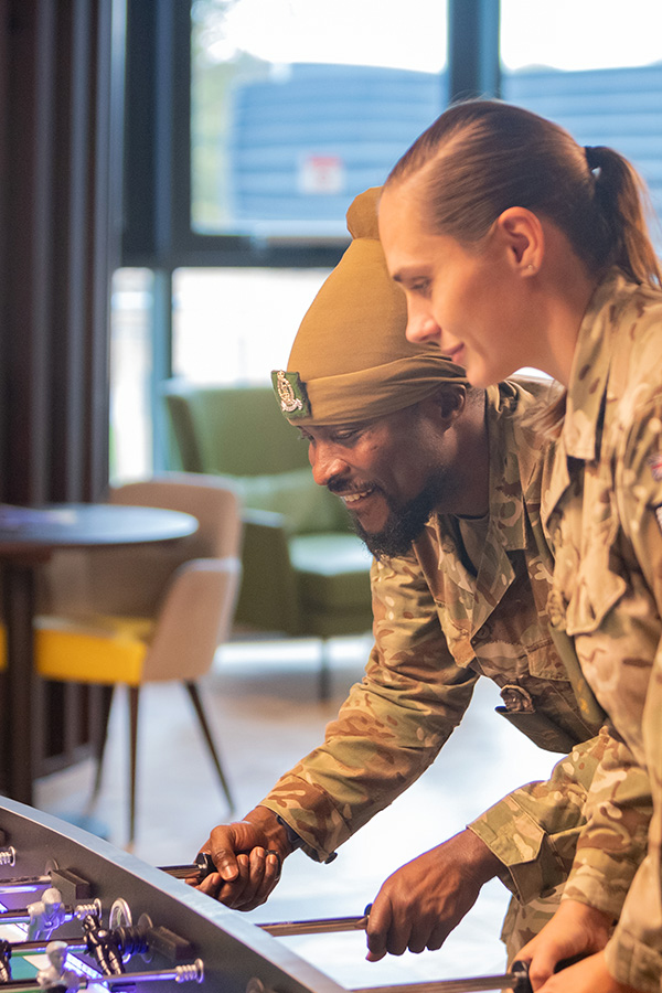 Two people in military uniforms play foosball indoors, smiling and focused, with a modern lounge area in the background.