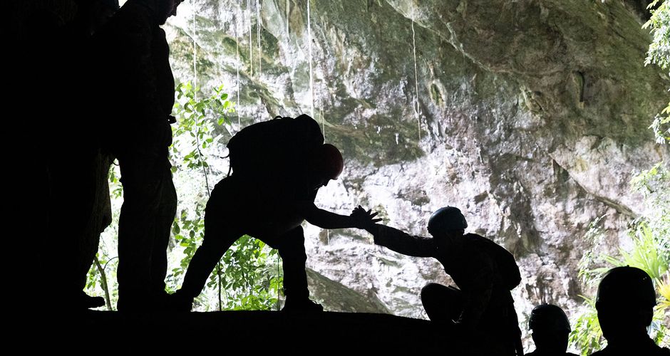 Silhouettes of people inside a cave, with one person helping another climb up, against a backdrop of rocks and greenery outside.