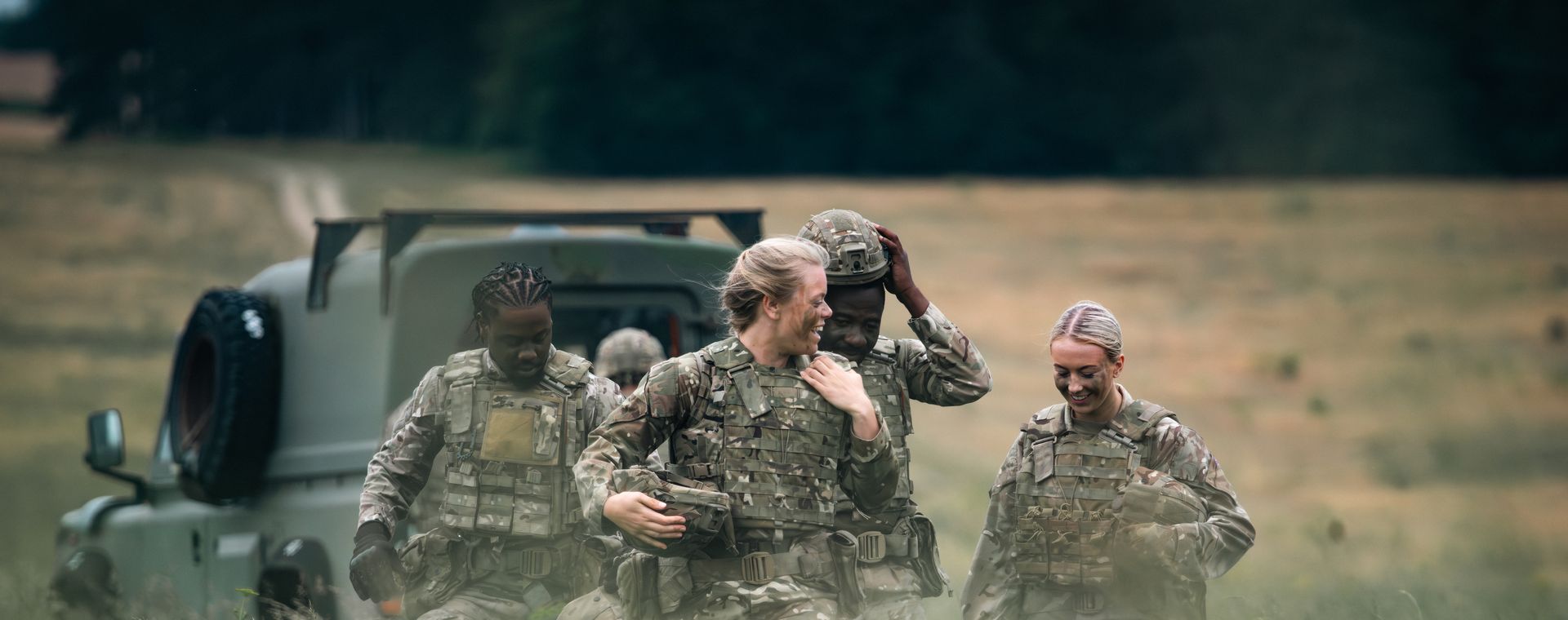 A group of soldiers in camouflage uniforms walk together through a grassy field, laughing and chatting near a military vehicle.
