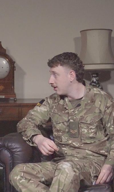Two people in military uniforms sit on leather chairs, talking. A vintage clock and lamps are on a wooden table in the background.