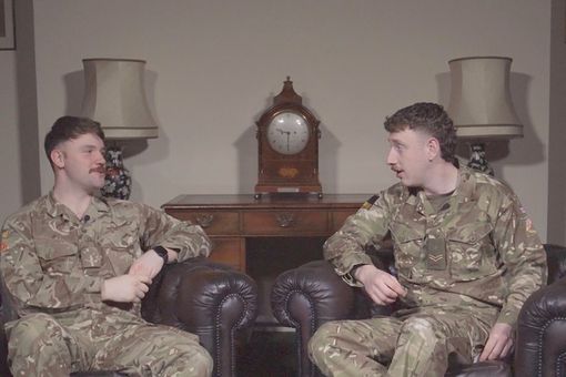 Two people in military uniforms sit on leather chairs, talking. A vintage clock and lamps are on a wooden table in the background.