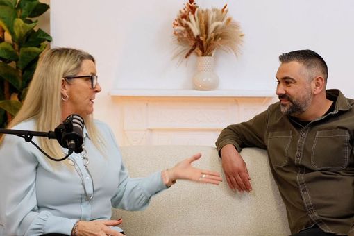 Two people sit on a couch having a conversation, with a microphone visible; plants and a vase are in the background.
