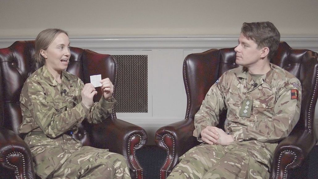 Two people in military uniforms sit in leather chairs, one holding a small card, engaged in conversation in a room with a radiator.