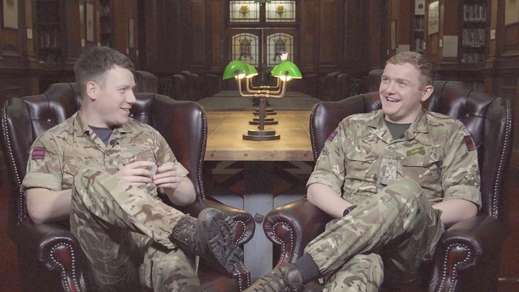 Two people in military uniforms sit in leather chairs, talking and smiling, in a wood-paneled room with a large table and green lamps.
