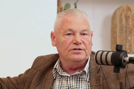 Man with short white hair and plaid shirt speaking into a microphone, seated indoors with decorative items in the background.