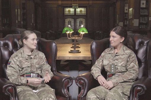 Two military personnel in uniform sit in leather chairs, engaged in conversation, in a dimly lit room with green lamps and wooden decor.