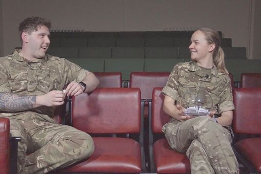 Two people in military uniforms sit on red chairs, smiling at each other in a dimly lit room. One holds a clear container.