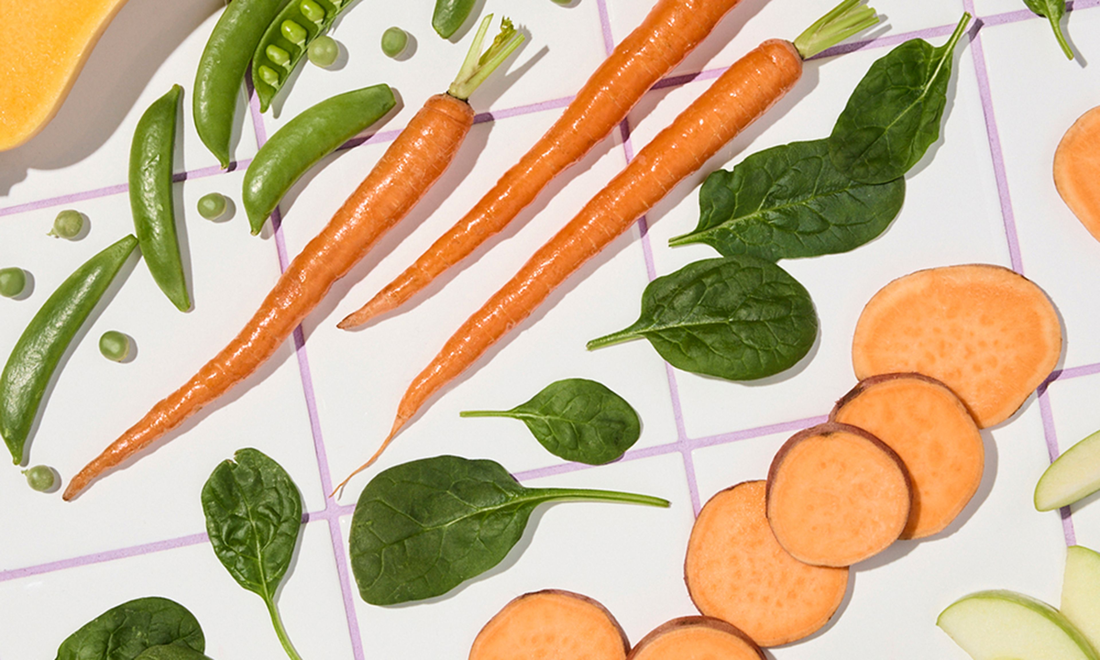 Assorted vegetables including carrots, snap peas, spinach leaves, and sliced sweet potatoes arranged on a white grid-patterned surface.