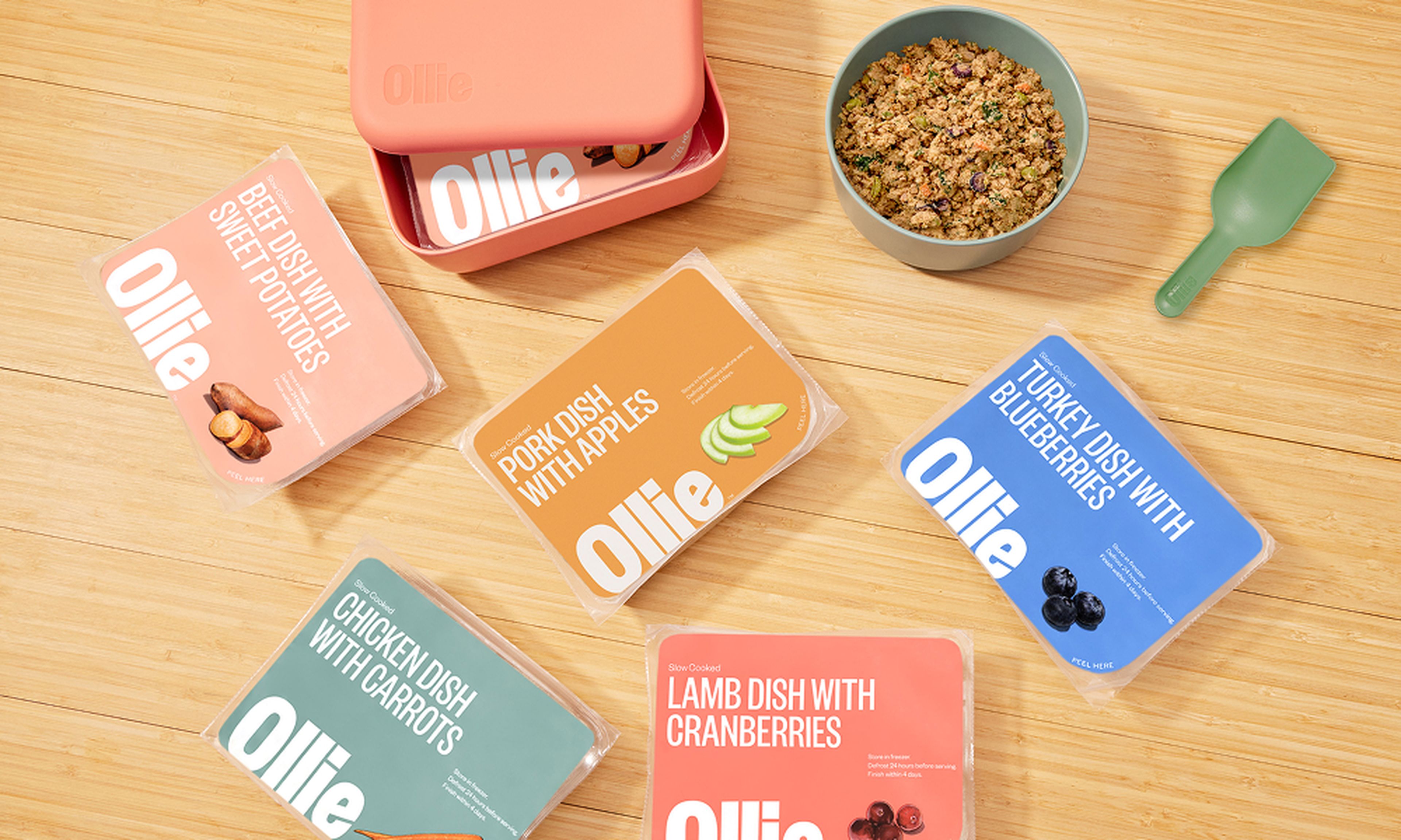 Assorted Ollie pet food packages and a bowl of mixed kibble on a wooden surface, with a small green scoop nearby.