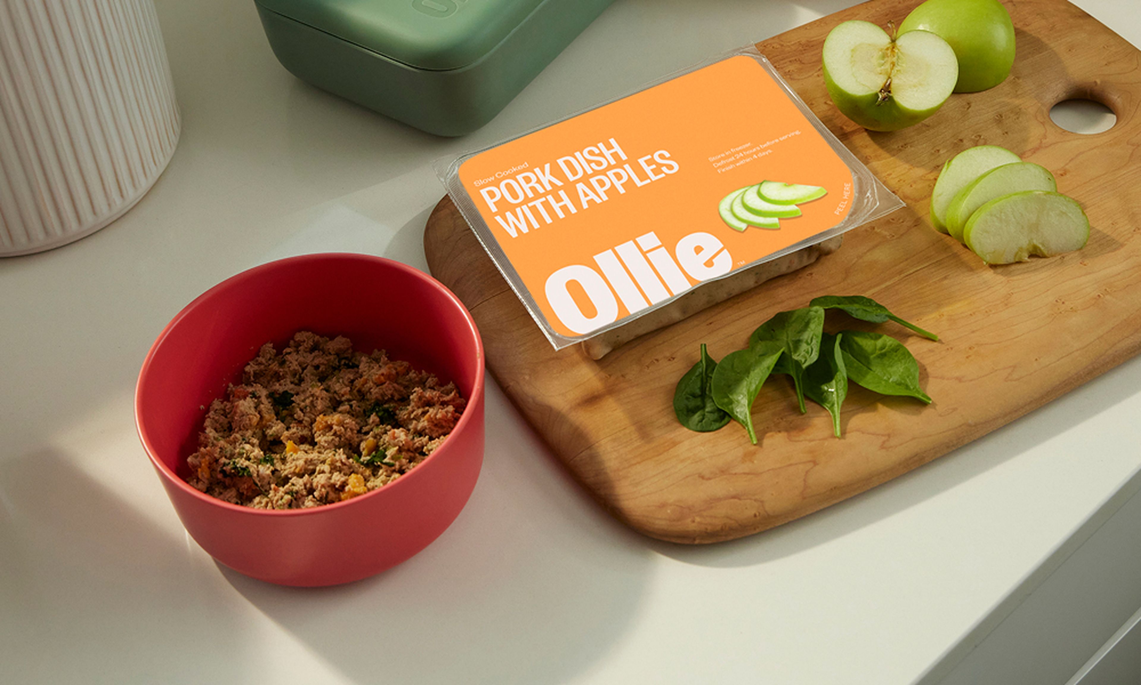A red bowl with pet food beside a wooden board holding a packaged pork dish with apples, spinach leaves, and sliced green apple.