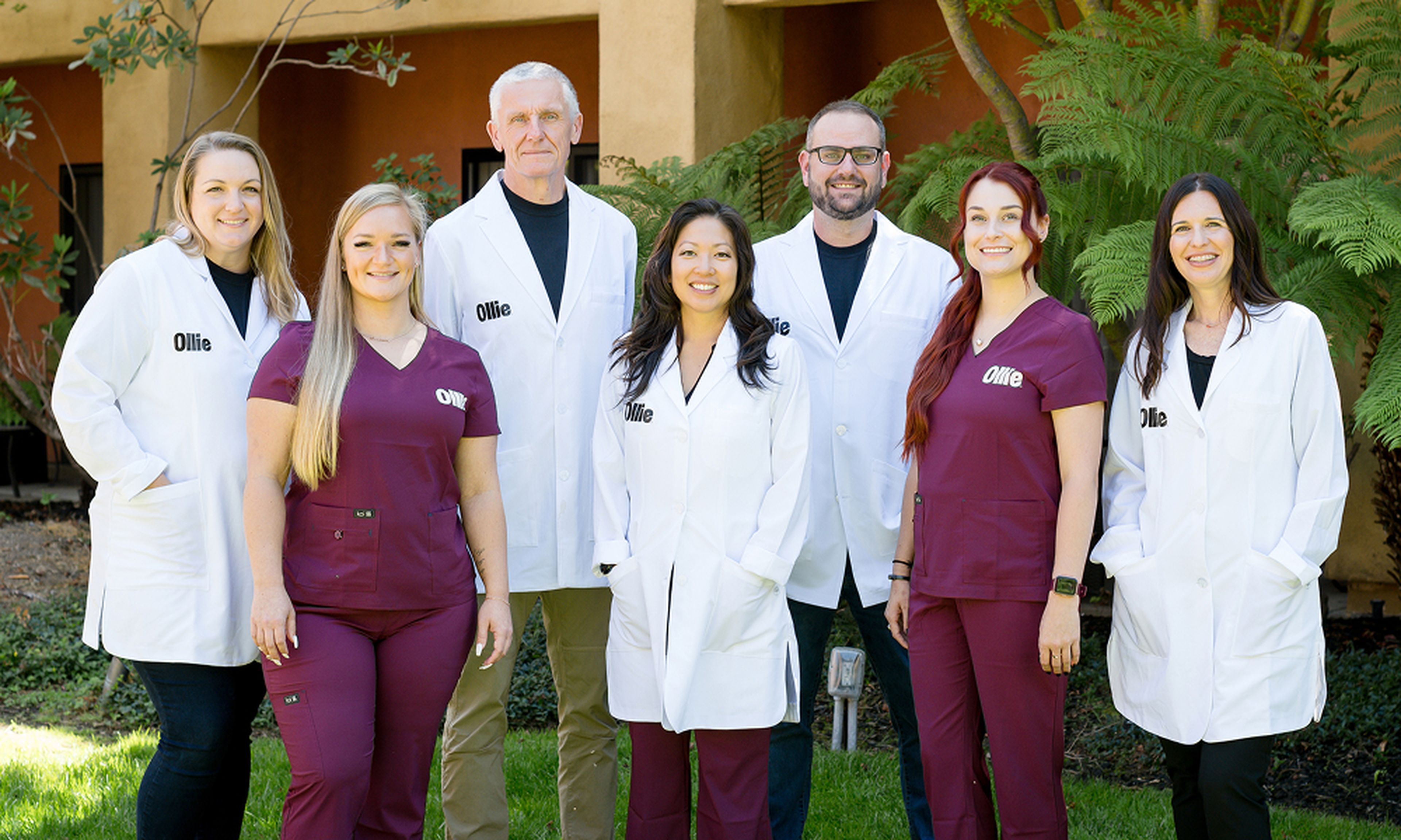 A group of seven people, some in white coats and others in maroon scrubs, standing outdoors in front of a building with greenery.