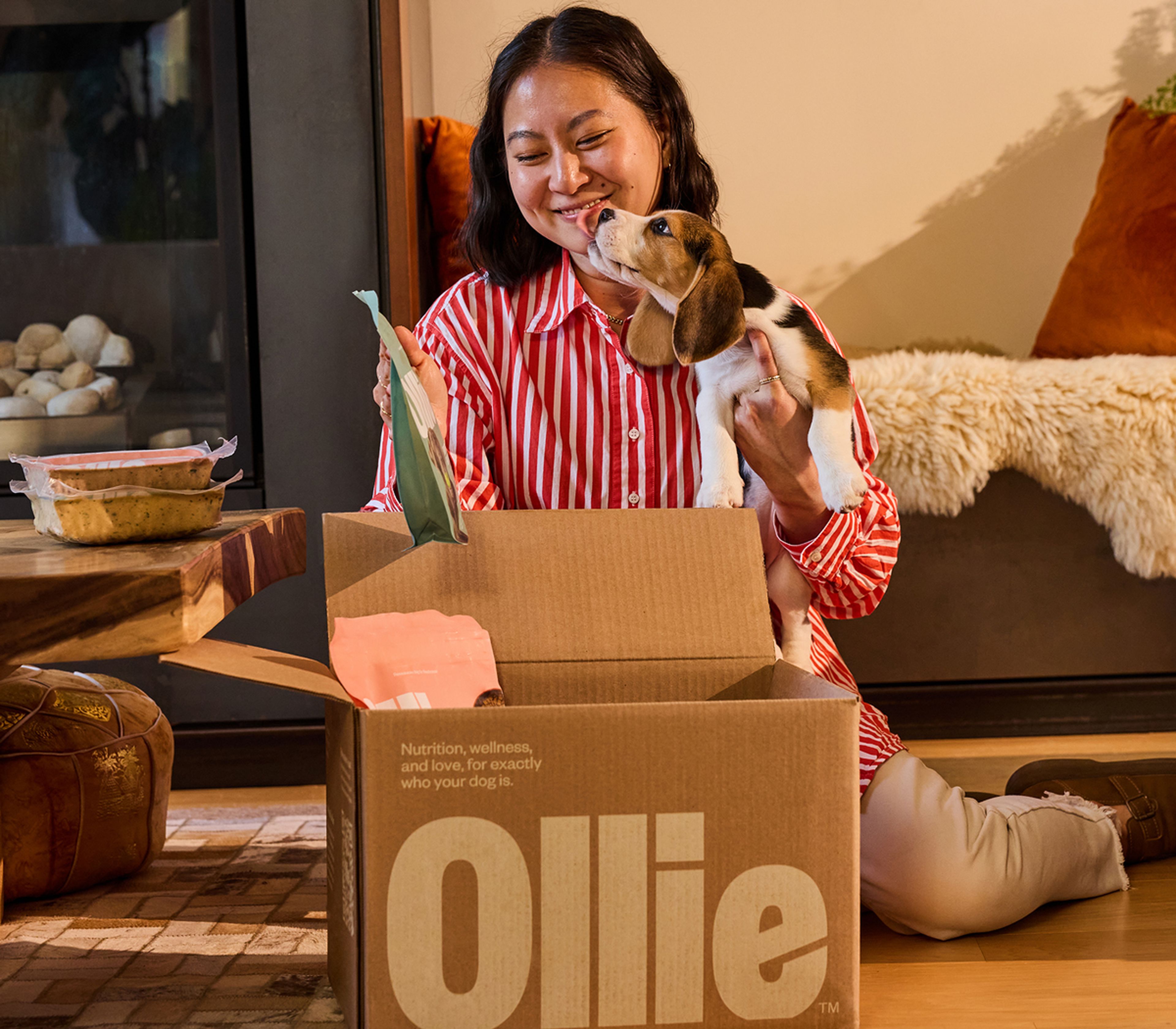 Image of a woman wearing a striped shirt holding a small dog that is licking her face while exploring the contents of her first Ollie delivery