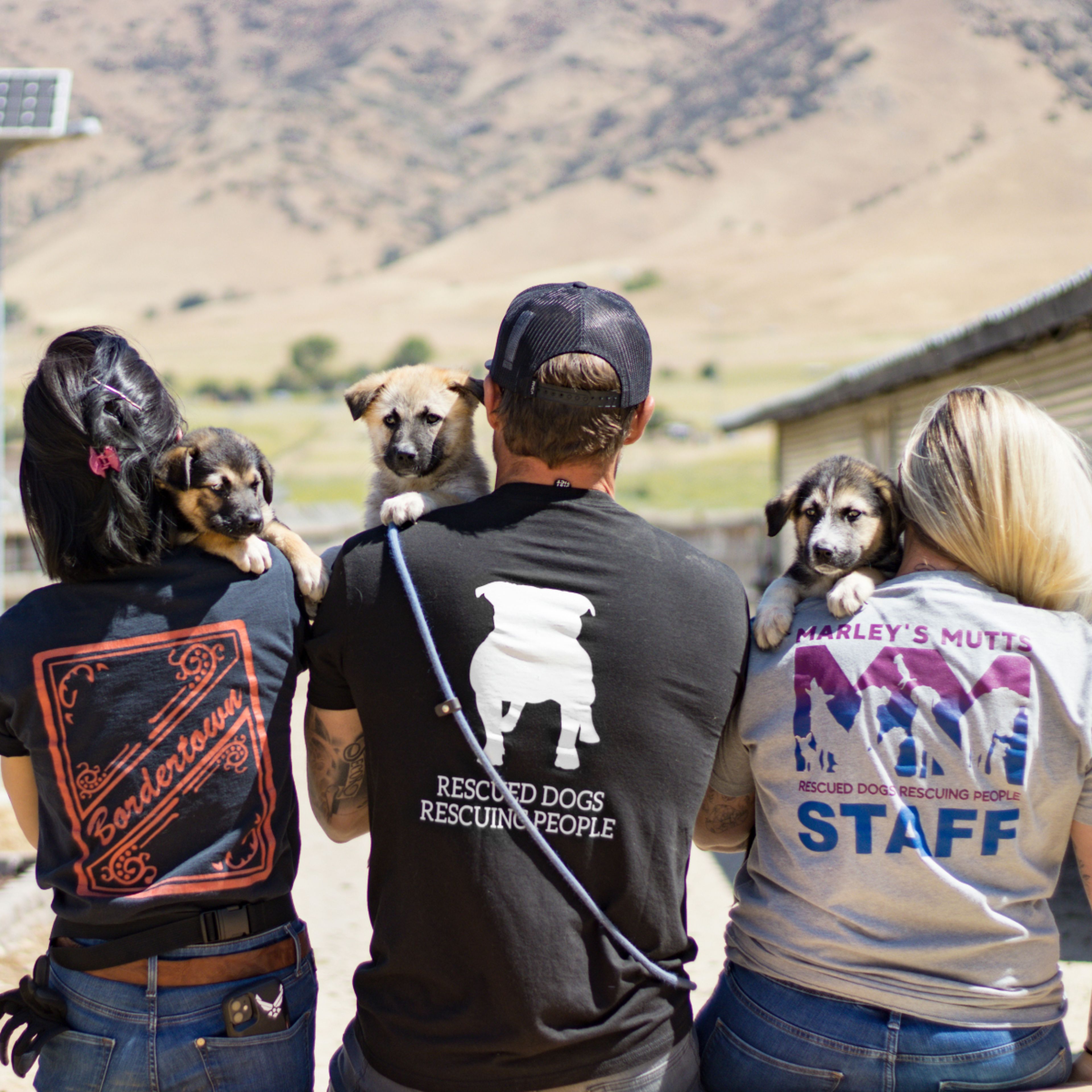 Four people with puppies on their shoulders stand outdoors, wearing shirts with rescue-themed designs. Hills and a barn are in the background.