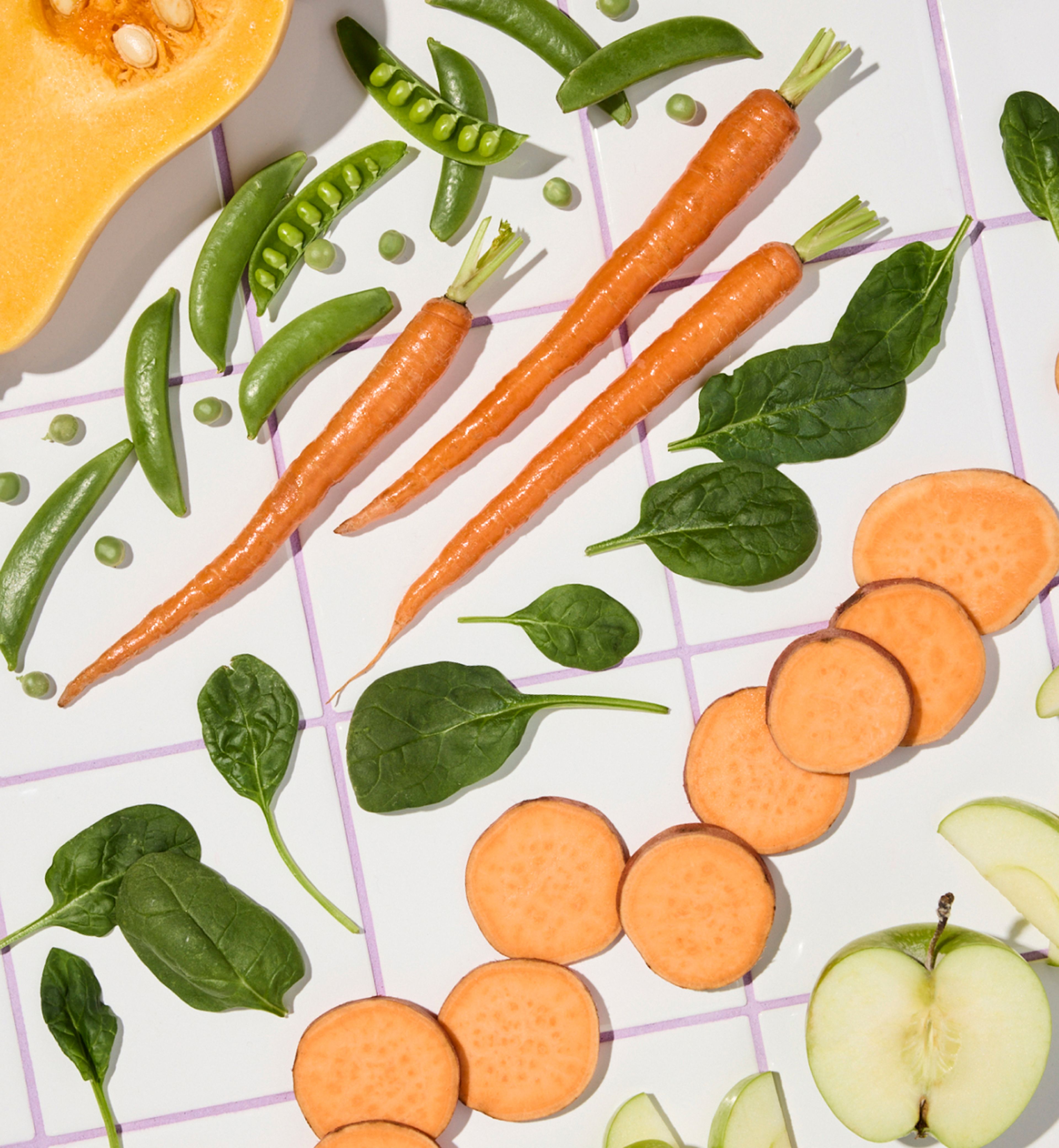 Assorted vegetables and fruits, including carrots, sweet potato slices, spinach leaves, snap peas, and apple slices, arranged on a tiled surface.