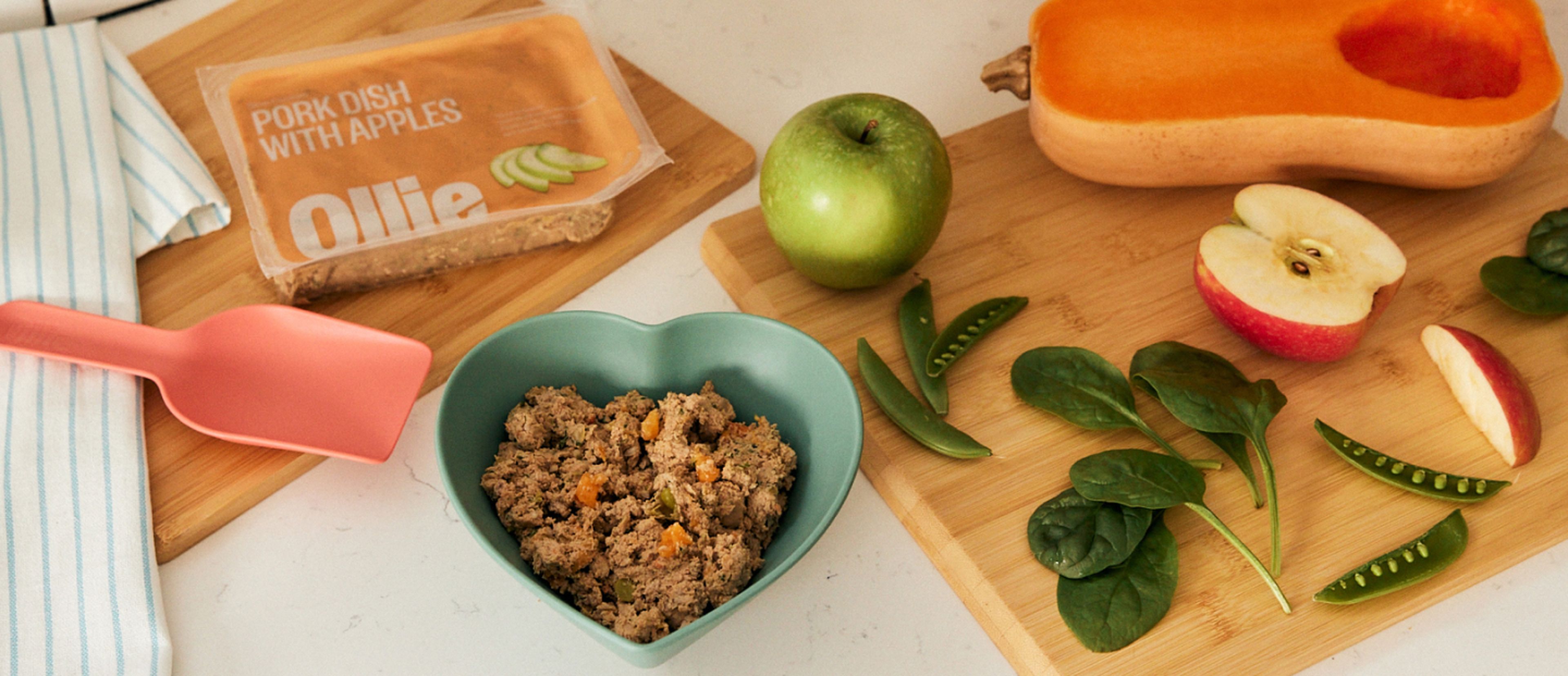 image of a cutting board with fresh fruits and veggies along side a heart shaped bowl of our fresh dog food.