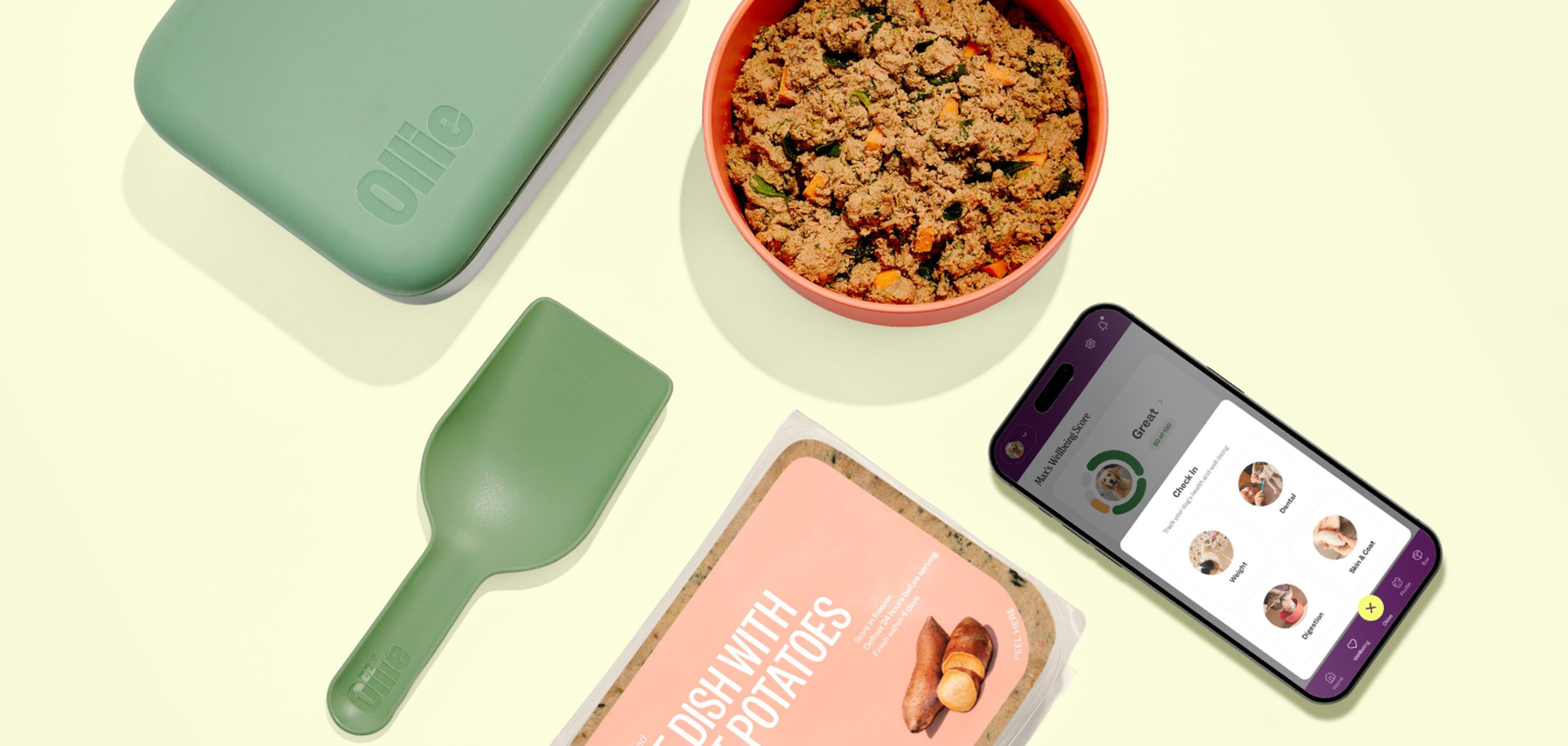 Dog food set with a green container, scoop, dish of food, phone with app displaying wellness score, and packaged dish with potatoes label.