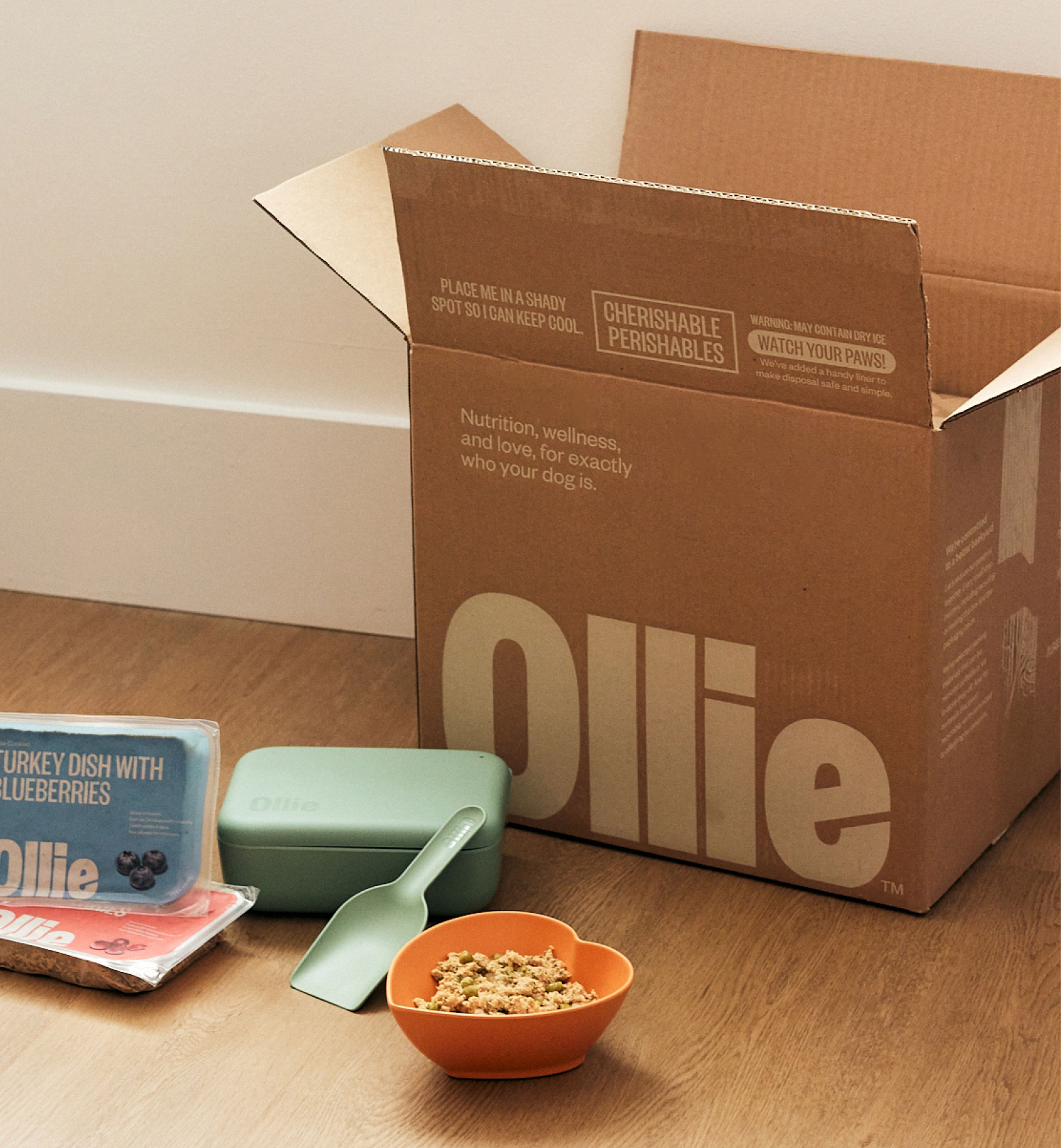 Image of an ollie box surrounded but Ollie fresh dog food packs, a hear shaped bowl and a scoop