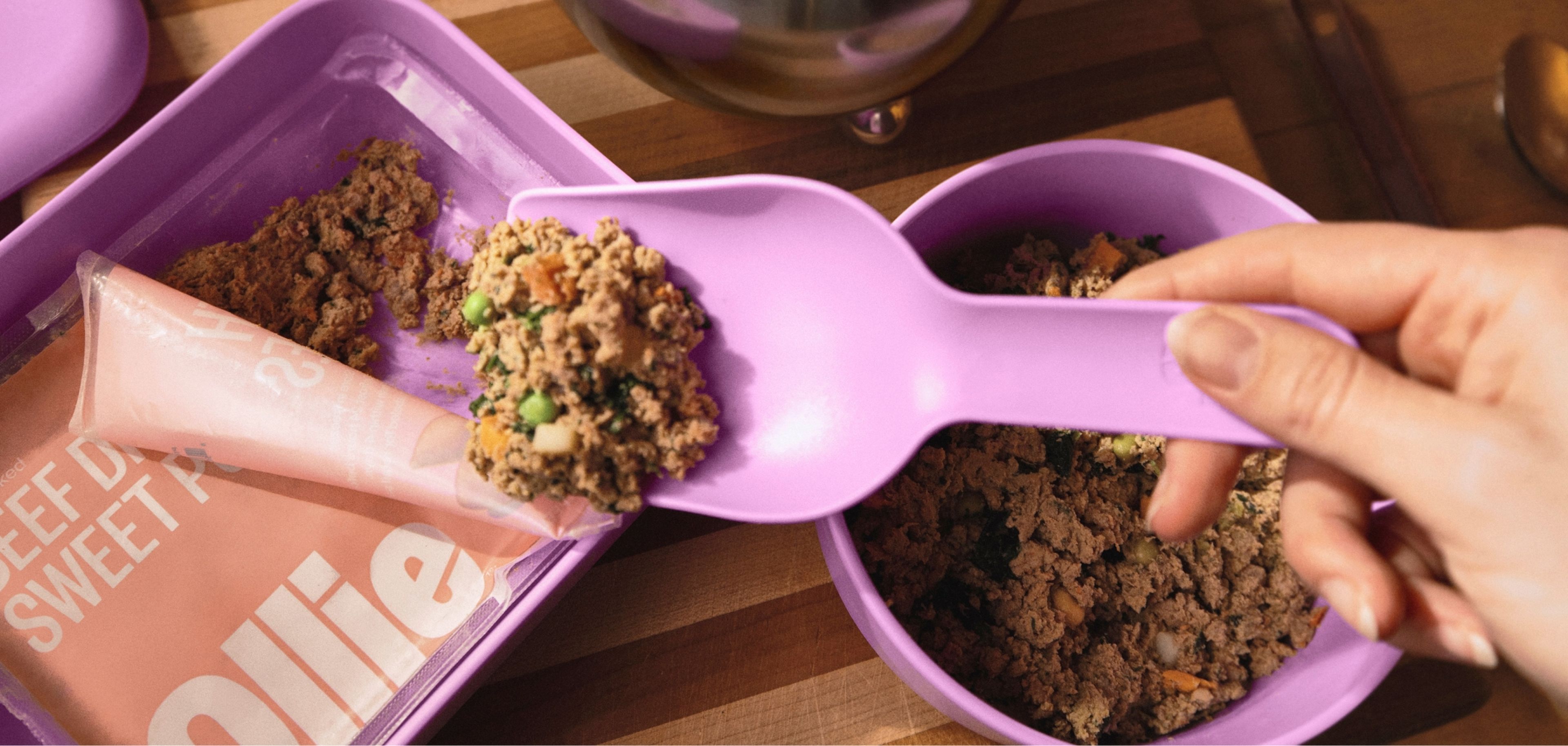 A hand uses a purple spoon to serve dog food from a tray into a purple bowl. A sachet labeled "Beef" is partially visible.