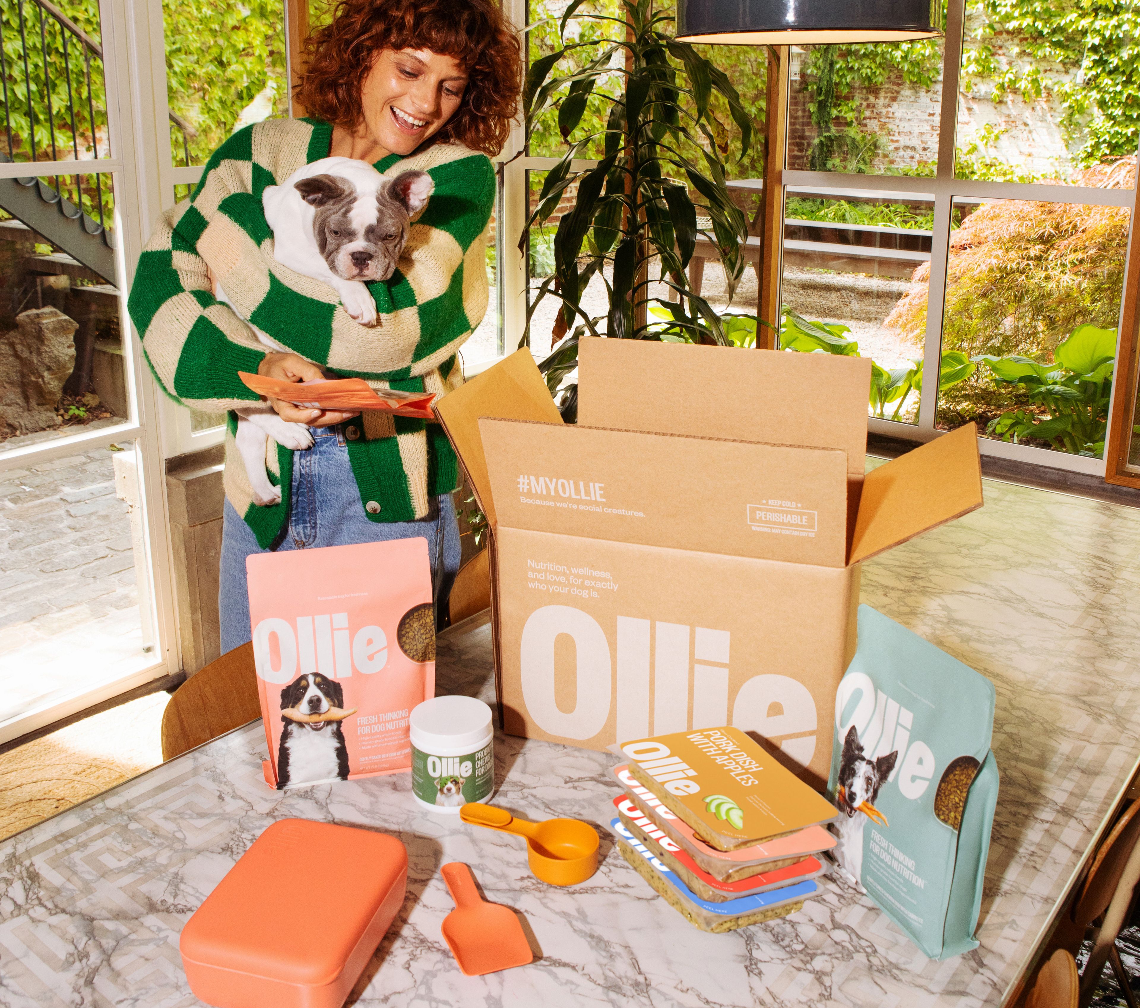 A woman holds a puppy next to a table with Ollie pet food products and an open box in a bright room with plants.